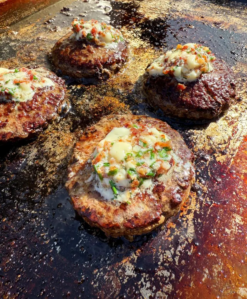 Four juicy burger patties cooking on a grill, each topped with melting compound butter mixed with herbs and small bacon bits, with sizzling juices and browned grill marks visible. This is a butter burger recipe