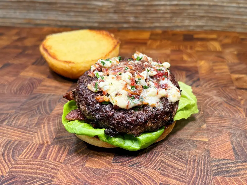 An open hamburger with a beef patty topped with melted cheese, herbs, and bits of bacon, resting on lettuce and a bottom bun. The top bun is placed nearby on a wooden surface.