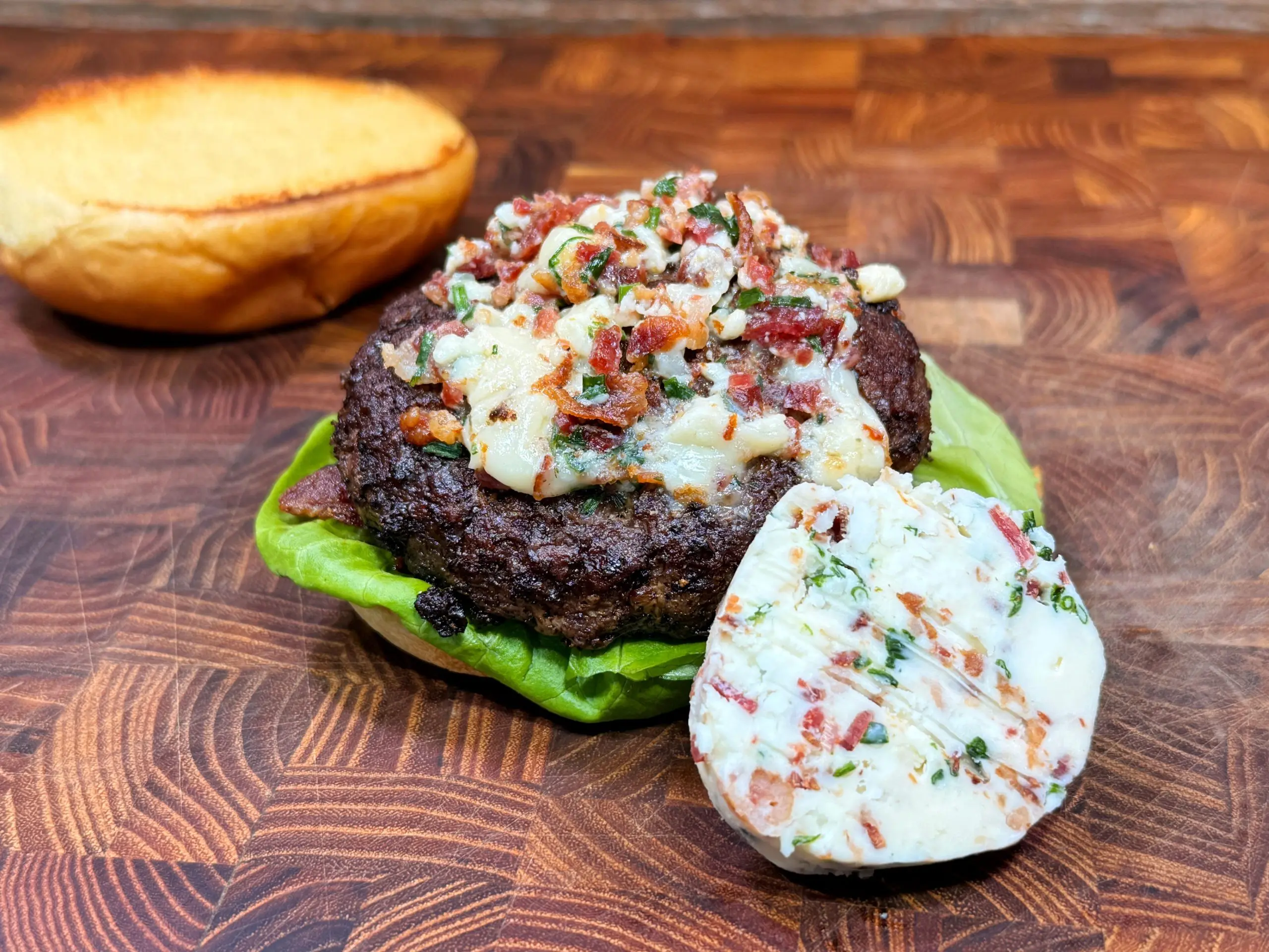 A cooked hamburger patty topped with chopped herbs and cheese sits on a leaf of lettuce, next to a slice of compound butter. A toasted top bun rests in the background on a wooden surface.