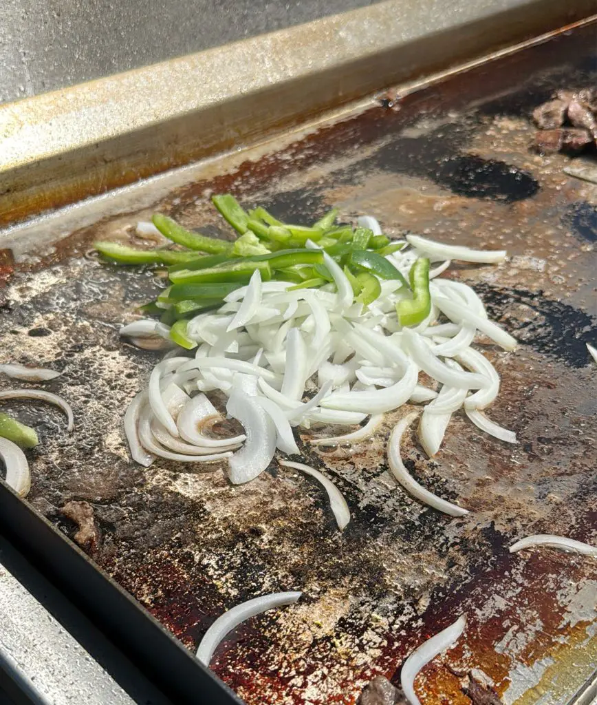 Sliced onions and green bell peppers are cooking on a flat top grill, with some browning and residue visible on the hot surface under bright sunlight.