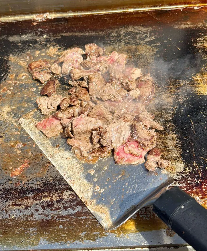 Chunks of beef cooking on a hot, greasy griddle, with some pieces browned and others still slightly pink. A large metal spatula with a black handle is lifting the meat, and steam rises from the sizzling surface.