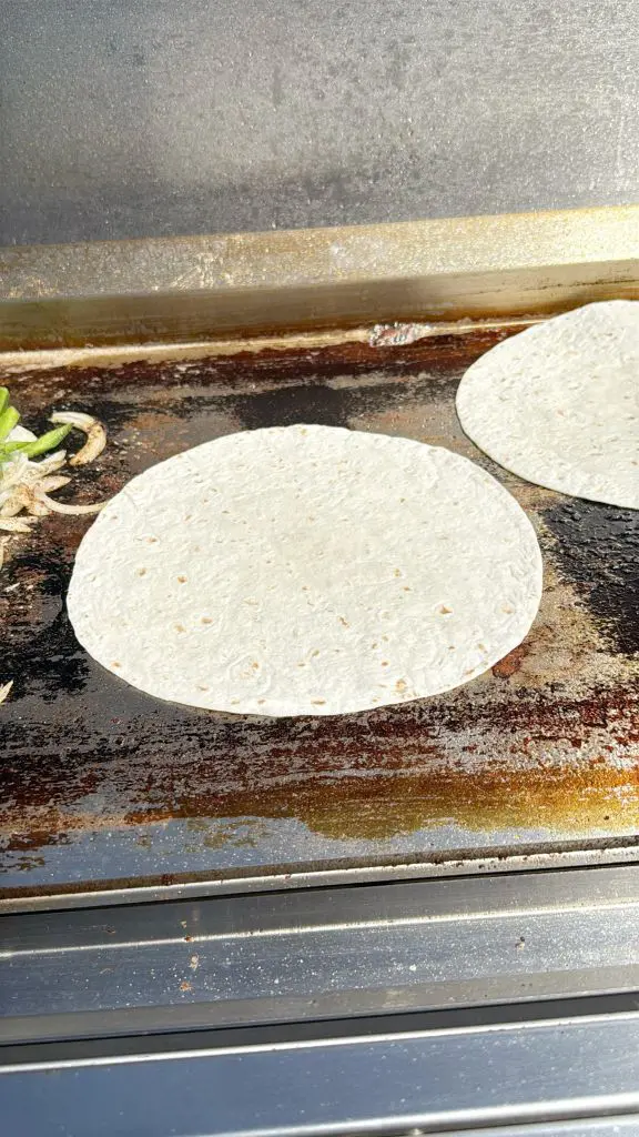 A flour tortilla is cooking on a hot, slightly greasy griddle. Some onions and vegetables are visible on the left side, with another tortilla in the upper right corner.