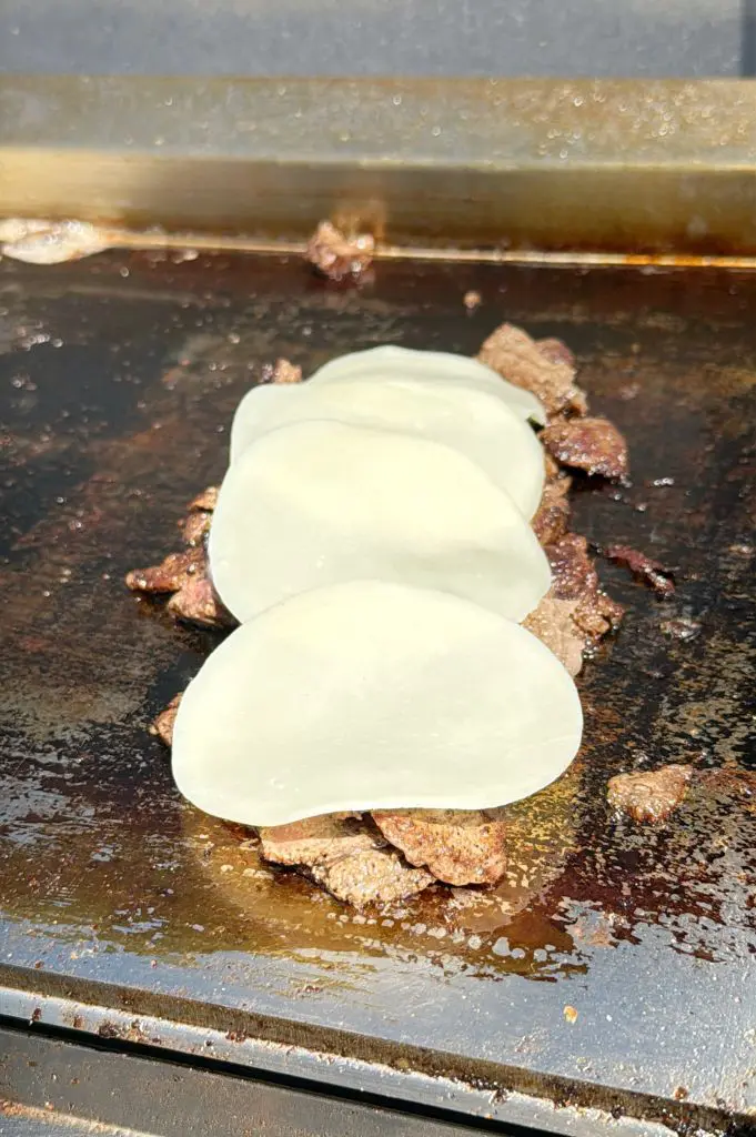 Thin slices of beef cooking on a flat griddle, topped with three overlapping slices of provolone cheese starting to melt.
