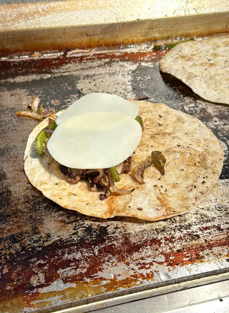 A tortilla filled with grilled beef, onions, and green peppers, topped with two slices of white cheese, cooking on a griddle beside another plain tortilla.