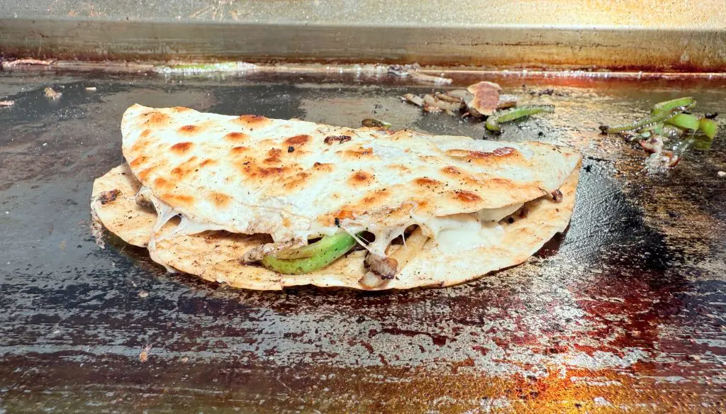 A toasted quesadilla filled with melted cheese, grilled meat, and green bell peppers sits on a hot, greasy griddle.