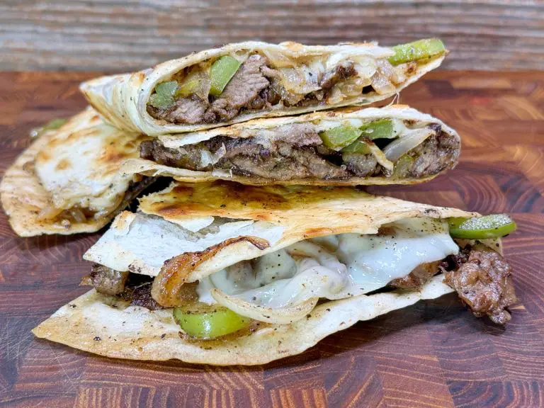 Three quesadilla wedges filled with sliced beef, melted cheese, onions, and green bell peppers are stacked on a wooden surface, showing the savory filling inside crisp, golden tortillas.