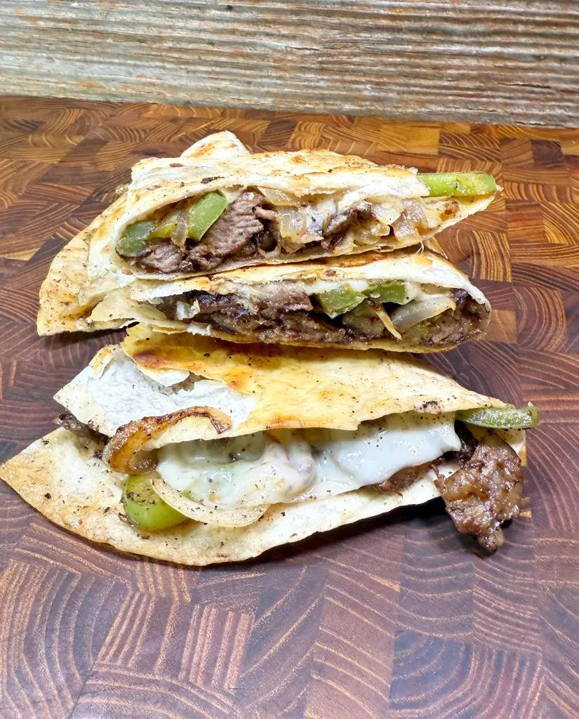 Three quesadillas filled with melted cheese, beef, onions, and green bell peppers are stacked on a wooden cutting board, with a rustic wooden background behind them.