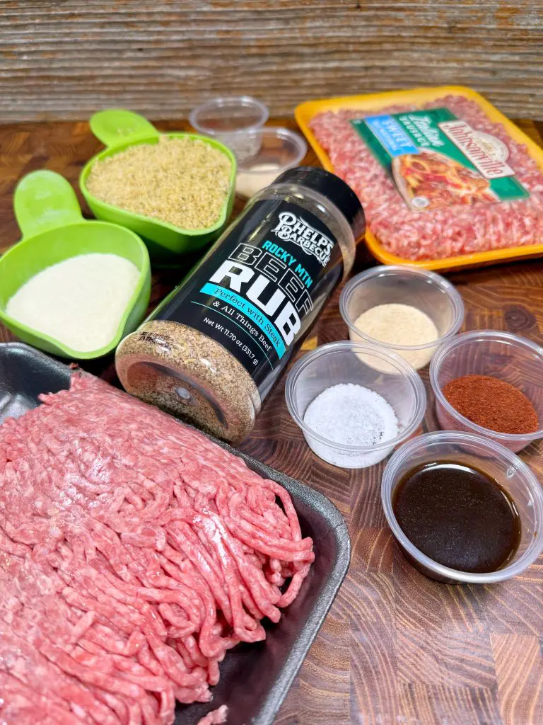 Ground beef, ground pork, green measuring cups with bread crumbs and milk, a container of beef rub seasoning, small bowls with salt, paprika, garlic powder, onion powder, and Worcestershire sauce on a wooden surface.