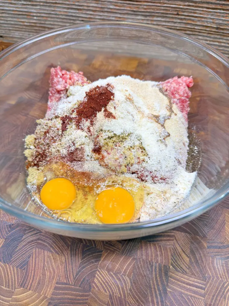 A glass bowl filled with raw ground meat, two cracked eggs, breadcrumbs, and various seasonings, resting on a wooden surface.