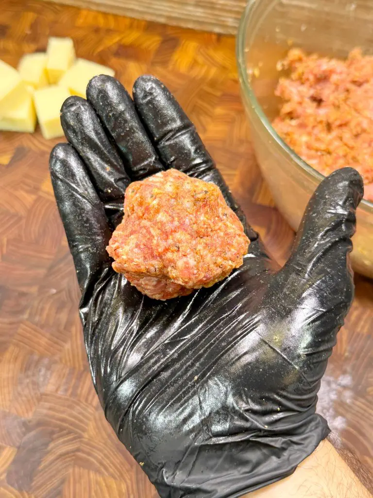 A hand wearing a black glove holds a raw meatball over a wooden surface. A glass bowl with more ground meat and some potato pieces are visible in the background.