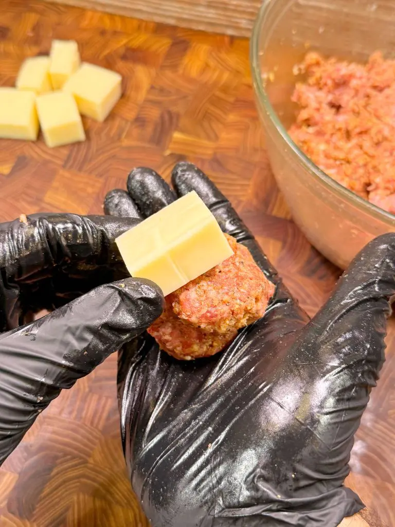 A person wearing black gloves holds a meatball and places a cube of cheese inside it, with more cheese cubes and a bowl of ground meat visible in the background on a wooden surface.