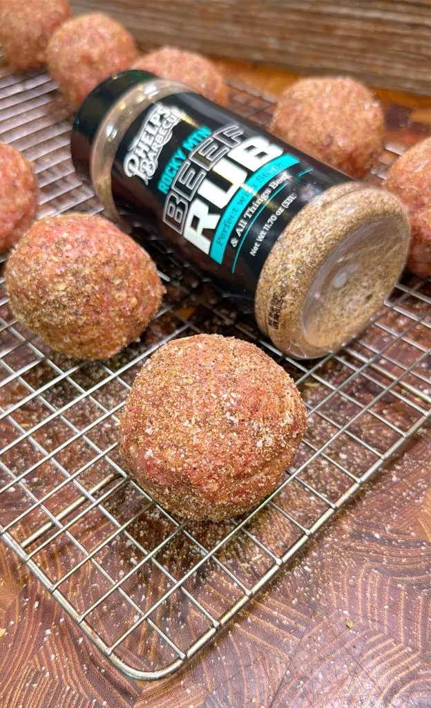 Seasoned meatballs on a wire rack, with a jar of Phelps BBQ Rocky MTN Beef Rub seasoning laying on its side, some seasoning spilled onto the rack and wooden surface.
