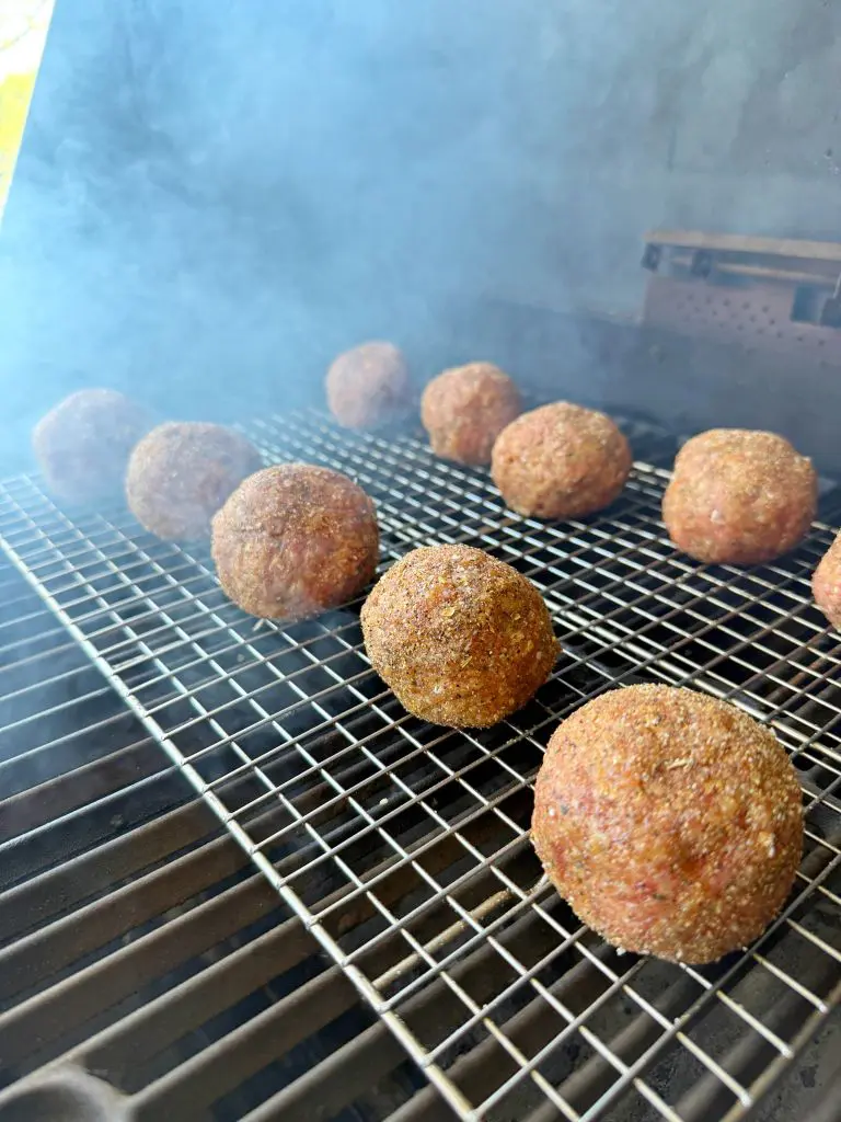 Meatballs coated in seasoning are arranged on a metal rack inside a smoker, with smoke rising around them as they cook.