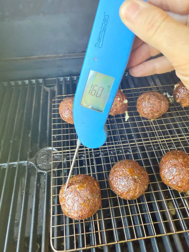 A hand holds a blue digital meat thermometer, showing 160°F, inserted into a cooked meatball on a wire rack inside a grill with several other meatballs.