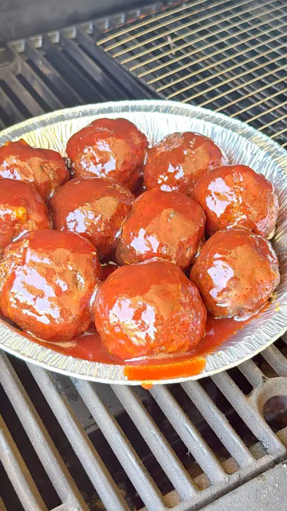 A foil pan filled with saucy barbecue meatballs sits on a grill, with the meatballs coated in a glossy red sauce. The grill grate is visible underneath and beside the pan.