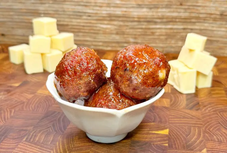 A white bowl holds three glazed meatballs, with cubes of cheese stacked in the background on a wooden surface.