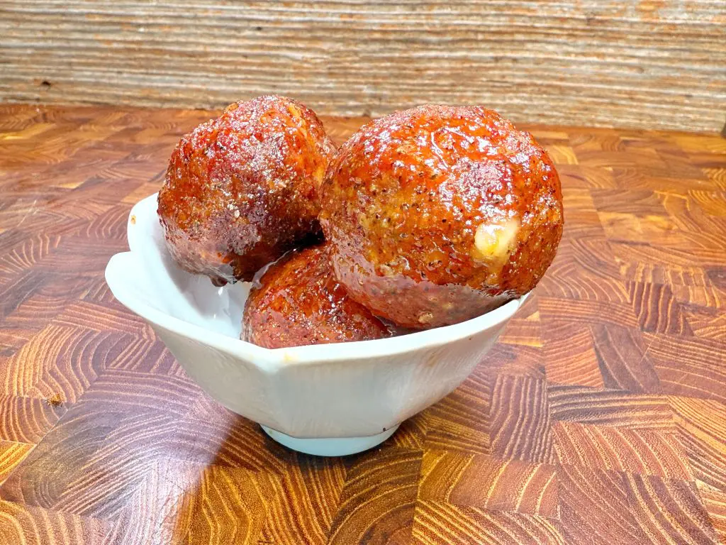 Three caramel-coated dessert balls are stacked in a small white bowl, set on a wooden surface with a patterned texture. The treats appear glossy and slightly sticky, with a golden-brown, sugary exterior.