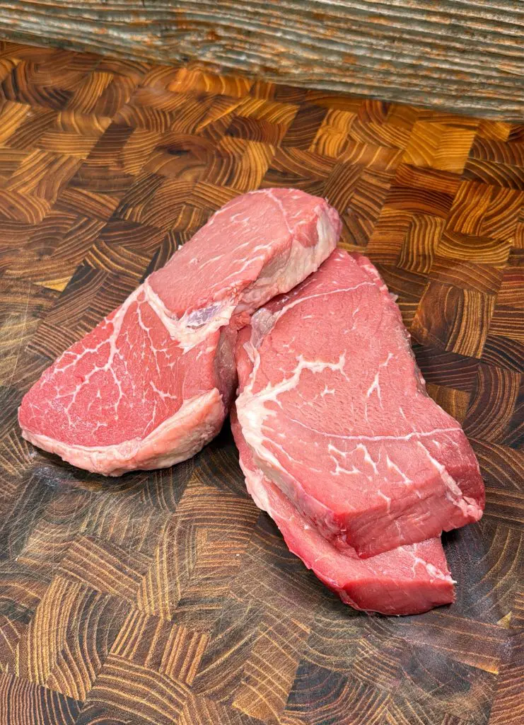 Three raw beef steaks with visible marbling are stacked on a wooden cutting board with a detailed grain pattern. The background includes a rustic, textured wooden surface.