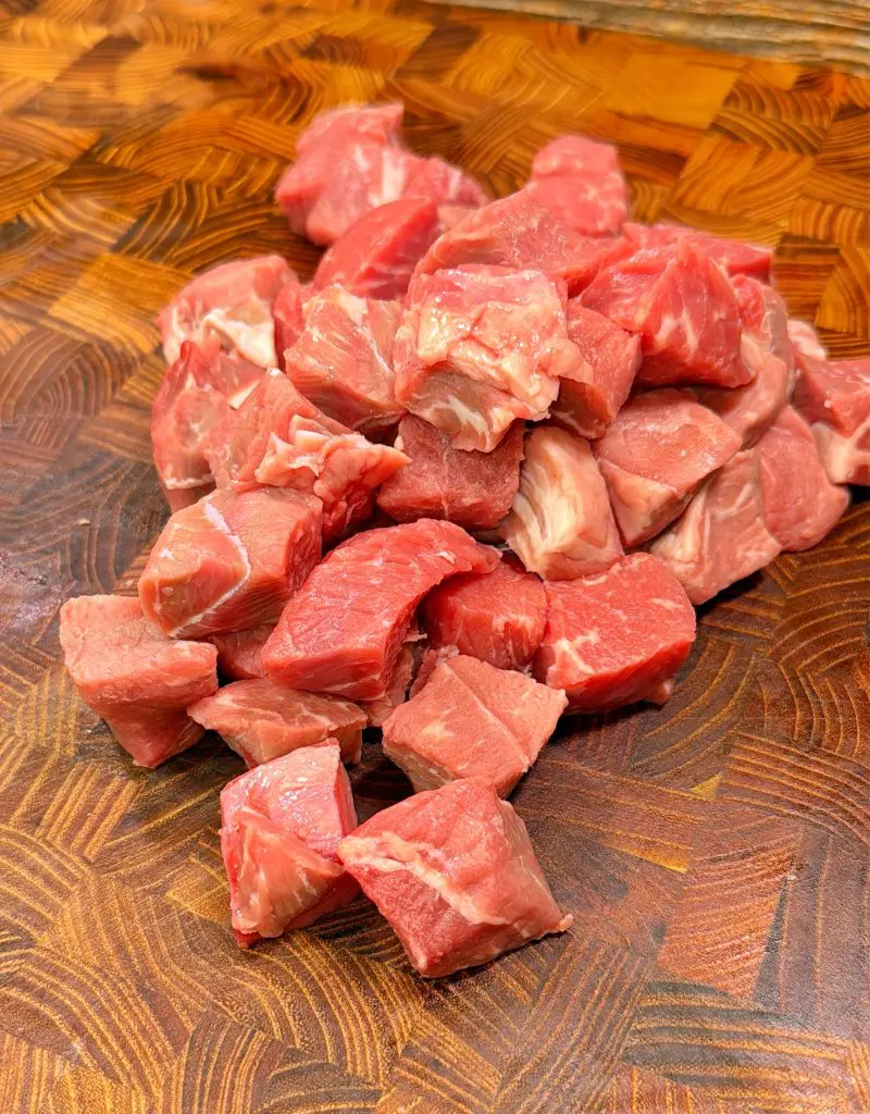 A pile of raw beef stew meat cut into cubes sits on a wooden cutting board with a geometric pattern.