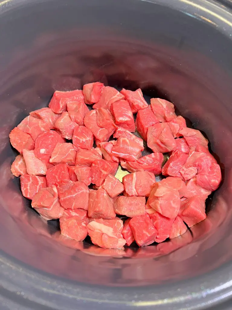 Cubed pieces of raw beef placed in the bottom of a black slow cooker, ready for cooking.