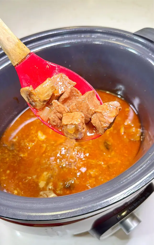 A red spoon holds chunks of cooked beef stew above a slow cooker filled with rich, brown stew broth and meat pieces.
