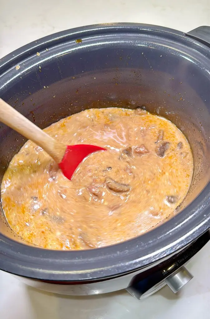 A red spatula stirs a creamy, orange-brown mixture with chunks of meat in a black slow cooker on a white countertop.
