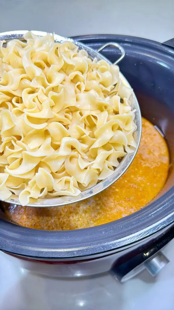 A metal strainer holds cooked egg noodles above a slow cooker filled with a creamy orange soup or sauce, ready to be combined with the crockpot steak tips.