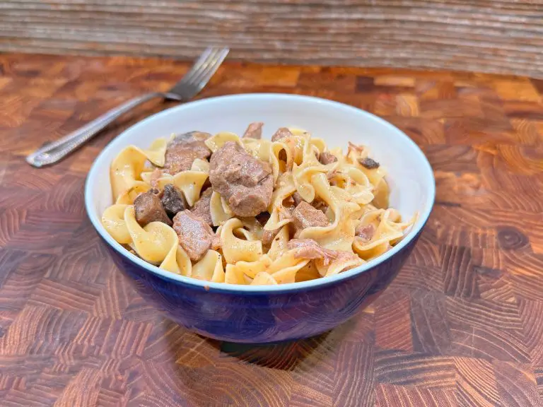 A bowl of beef stroganoff with egg noodles sits on a wooden countertop, with a fork in the background. The dish includes chunks of beef, mushrooms, and creamy sauce mixed with the noodles.