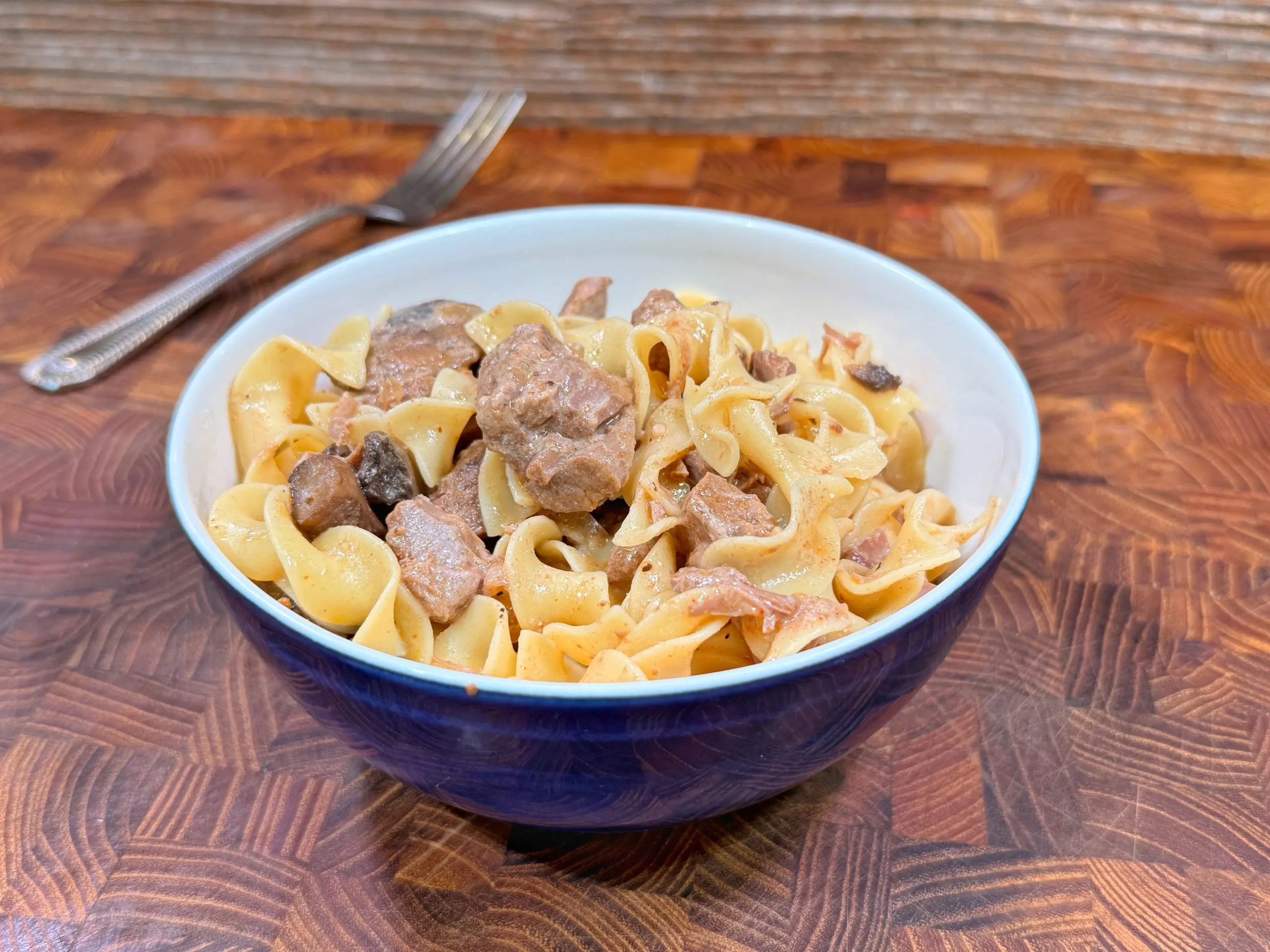 A blue and white bowl filled with egg noodles and chunks of beef sits on a wooden table, with a metal fork in the background.