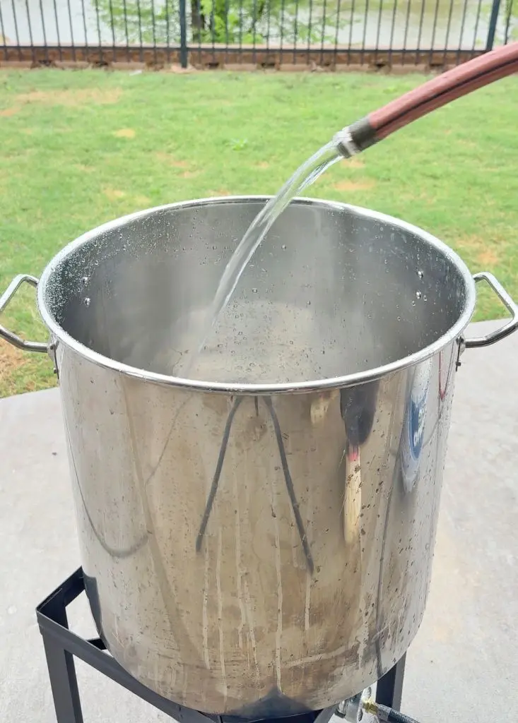 A large metal pot is being filled with water from a hose outdoors, placed on a propane burner on a concrete patio, with grass and a fence visible in the background.