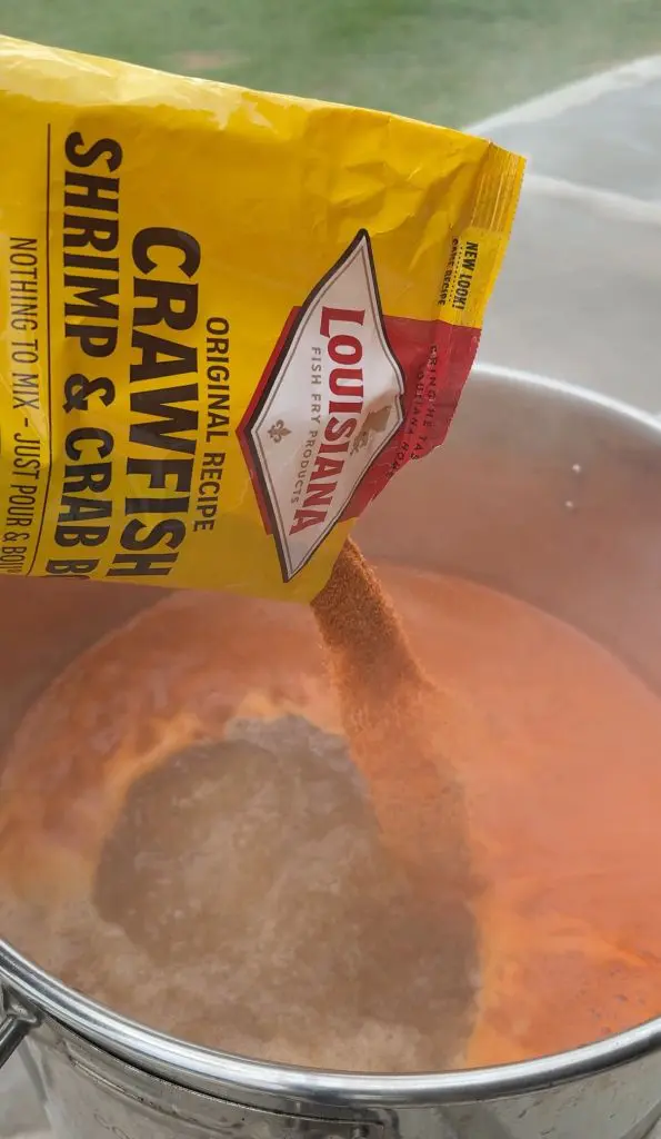 A hand pours Louisiana Crawfish, Shrimp & Crab Boil seasoning from a yellow packet into a steaming pot of boiling water.