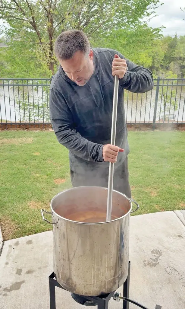 A man in a dark hoodie stirs a large steaming pot outdoors on a patio, with green grass, trees, and a lake visible in the background.