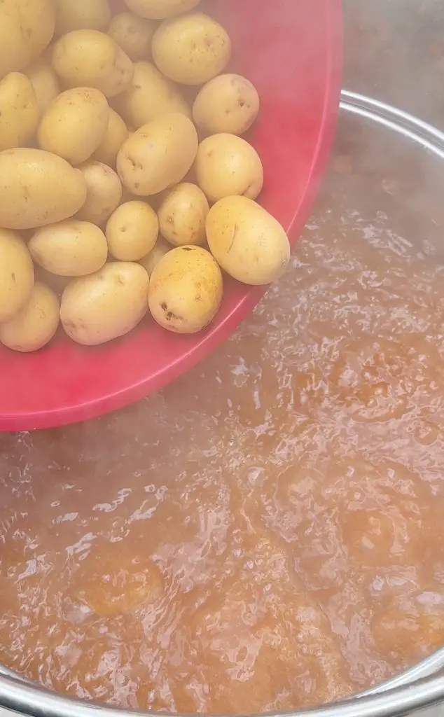 A red bowl of raw potatoes is being tilted over a large pot of boiling water, with steam rising up around the food.