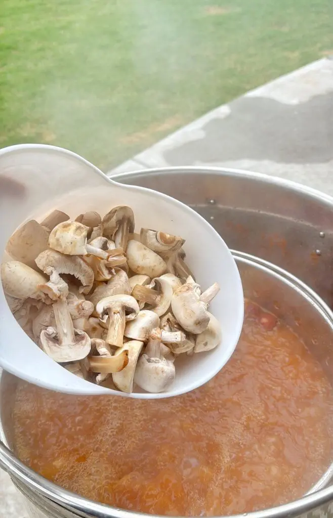 A white bowl of chopped mushrooms is being poured into a pot of bubbling orange soup or stew, outdoors on a patio with green grass visible in the background.