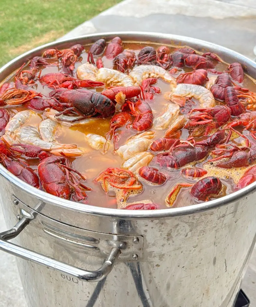 A large metal pot filled with boiling water, red crawfish, and seasonings, cooking outdoors on a bright day with grass visible in the background. This is an image of a crawfish boil recipe