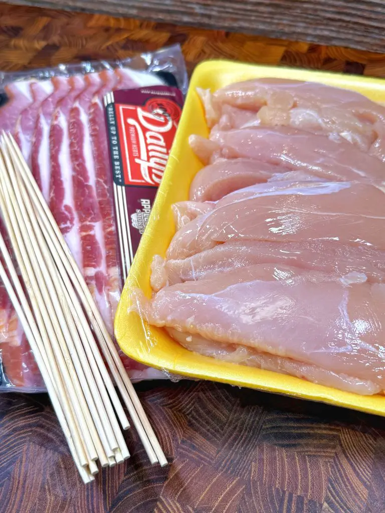 A package of raw bacon, a yellow tray of raw chicken breast fillets, and a bundle of wooden skewers arranged on a wooden surface. Ready to make chicken bacon ranch skewers