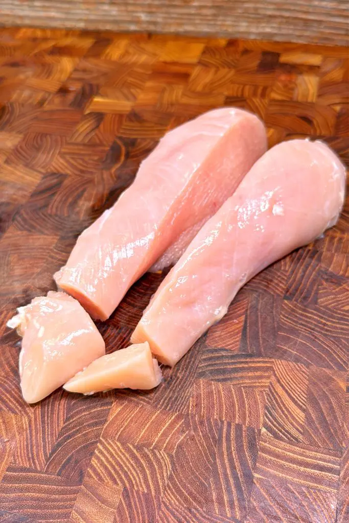 Two raw chicken breasts and two smaller pieces are placed on a wooden cutting board with a patterned grain.