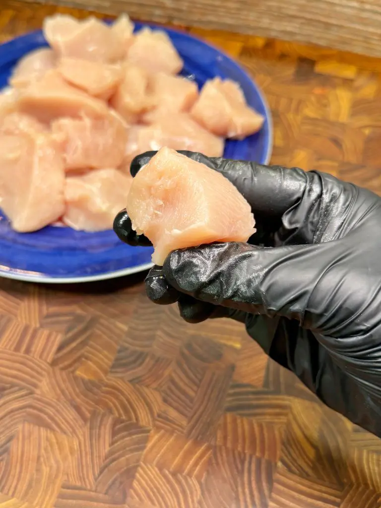 A hand wearing a black glove holds a piece of raw chicken over a wooden surface, with a blue plate containing more pieces of raw chicken in the background.