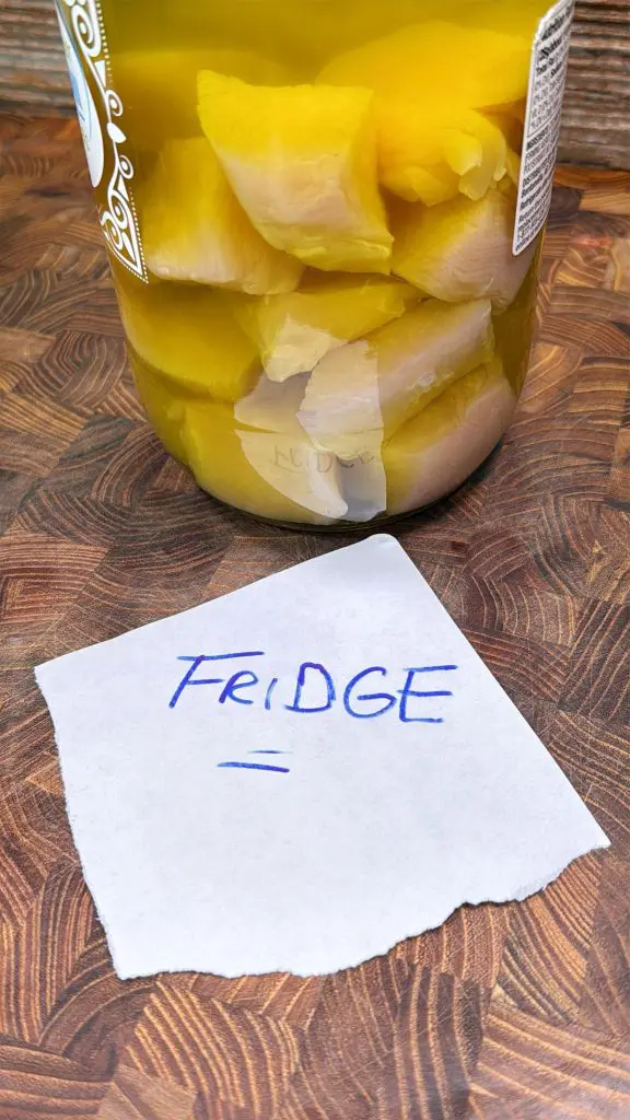 A jar filled with yellow pickled vegetables sits on a wooden surface. In front of the jar is a piece of paper with “FRIDGE =” handwritten in blue ink.