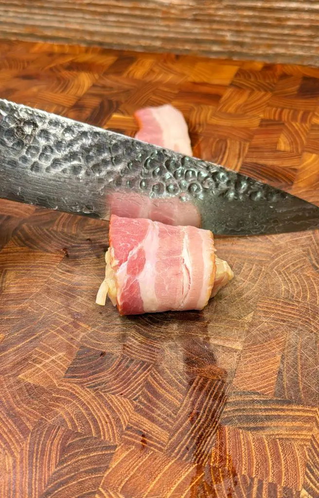A close-up of a knife slicing through a bacon-wrapped food item on a wooden cutting board.