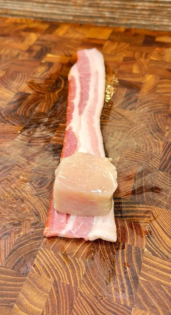 A raw scallop is placed on one end of a strip of uncooked bacon, set on a wooden cutting board with a geometric pattern.