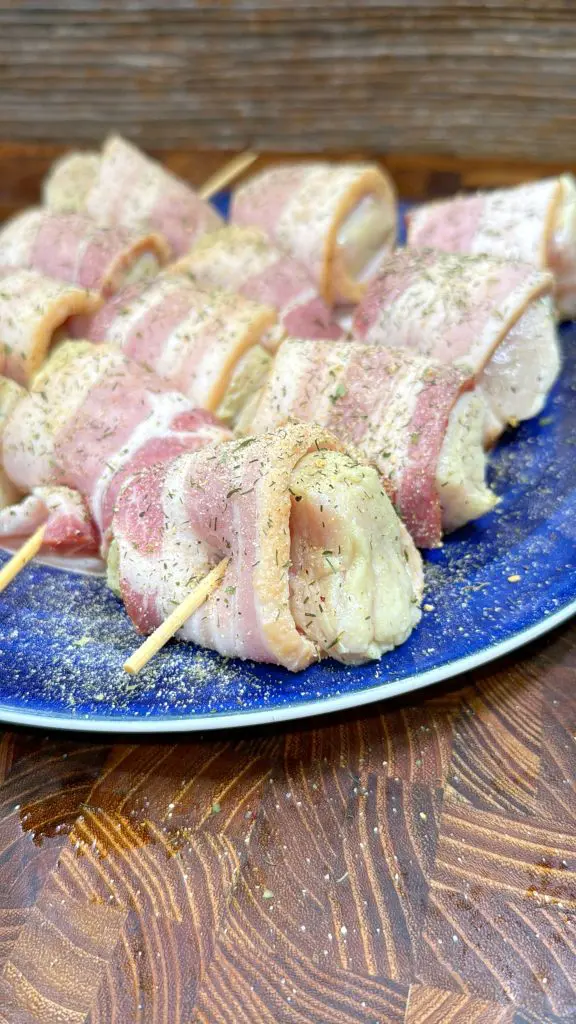 Raw bacon-wrapped chicken pieces secured with toothpicks, sprinkled with herbs and seasoning, arranged on a blue plate atop a wooden surface.