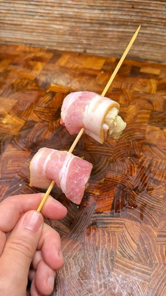 A hand holds a wooden skewer with two pieces of uncooked, bacon-wrapped food, likely chicken, on a wooden cutting board background.