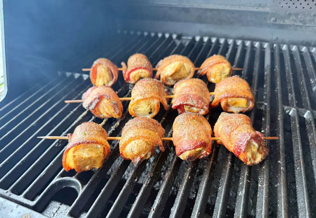 Bacon-wrapped pieces of meat on skewers are grilling on a barbecue, sizzling and browning over the heat with a smoky atmosphere visible in the background.