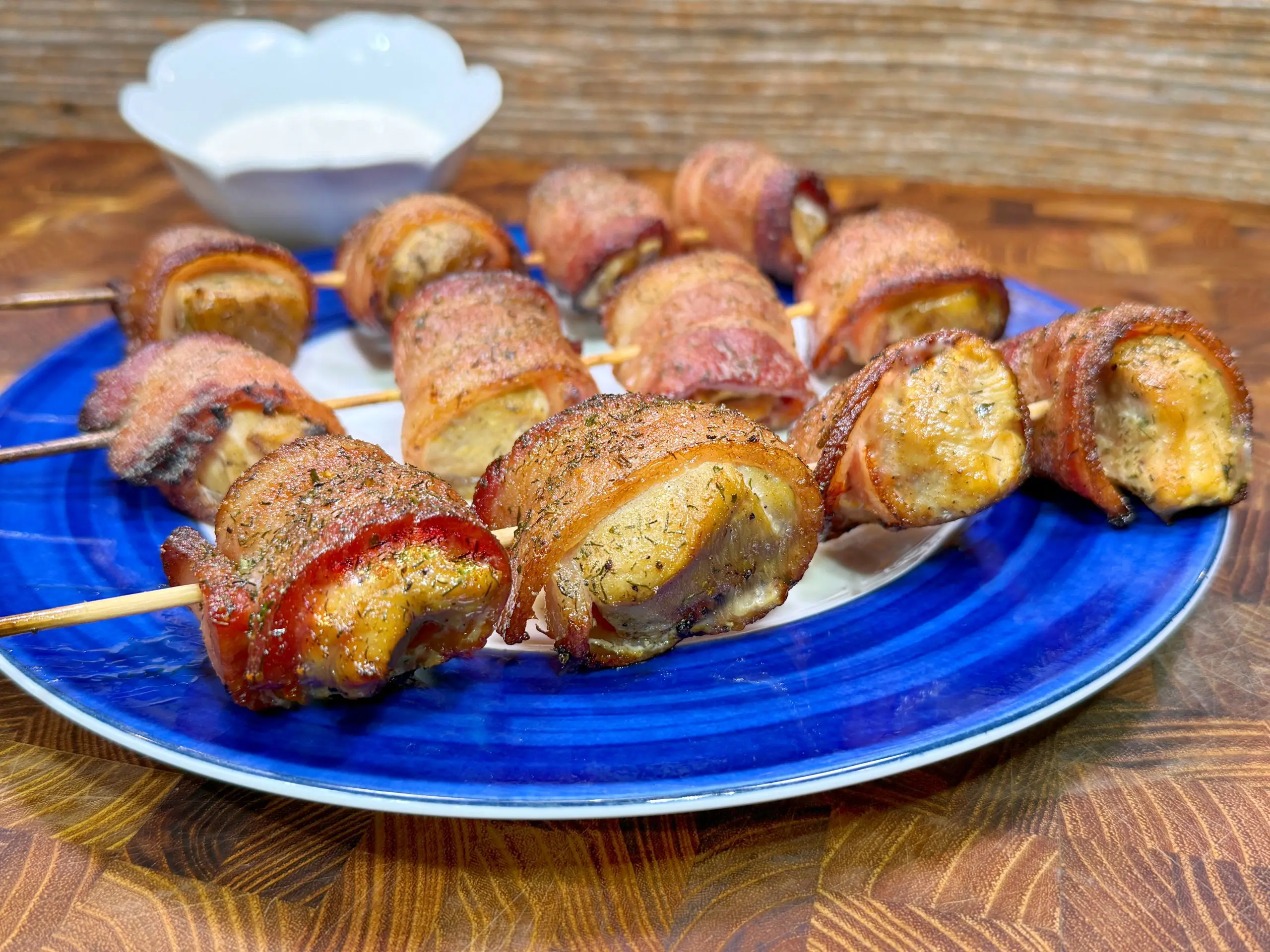 A blue plate holds skewered, bacon-wrapped chicken bites, seasoned and baked, with a bowl of white dipping sauce in the background on a wooden surface.