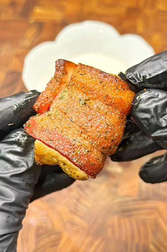 A close-up of hands in black gloves holding a seasoned, cooked piece of pork belly, with a blurred white bowl and wooden surface in the background.