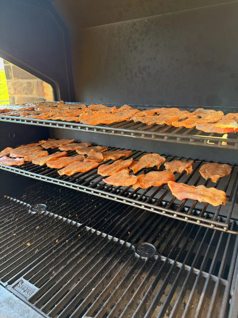 Sliced pieces of seasoned meat are spread out on two racks inside a smoker or grill, cooking slowly. The grill interior is clean and there is no food on the bottom rack.