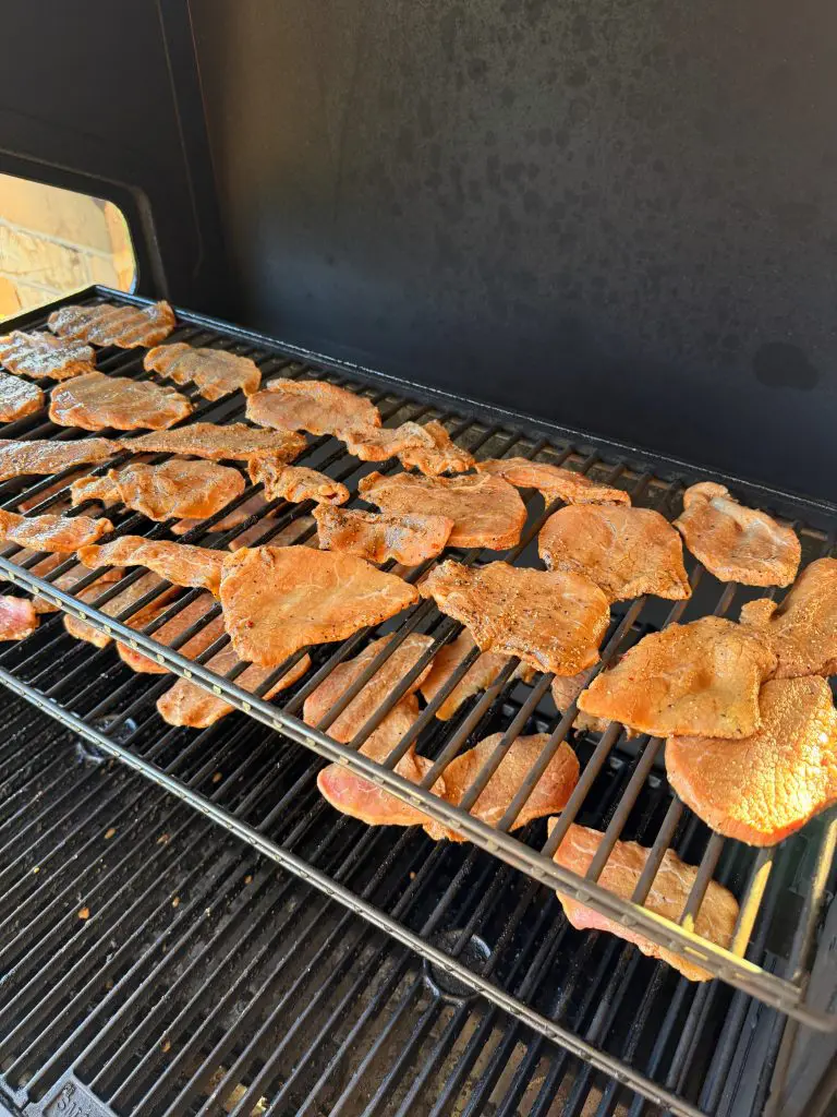 Thin slices of seasoned meat are arranged on the upper and lower racks of a black outdoor grill, cooking over the heat. Sunlight shines on the meat, highlighting its golden brown color.