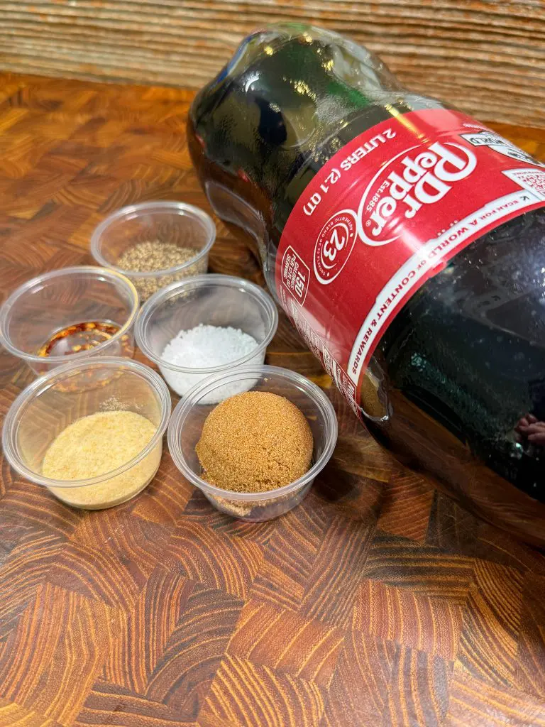 A bottle of Dr Pepper lies on its side next to six small containers holding various spices and seasonings, including brown sugar, salt, dried onion, and others, all arranged on a wooden surface.