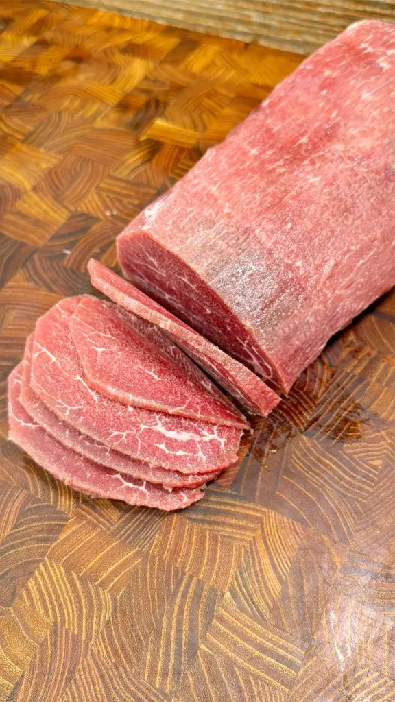 A log of raw beef is partially sliced on a patterned wooden cutting board, with several thin slices fanned out in front of the unsliced portion.